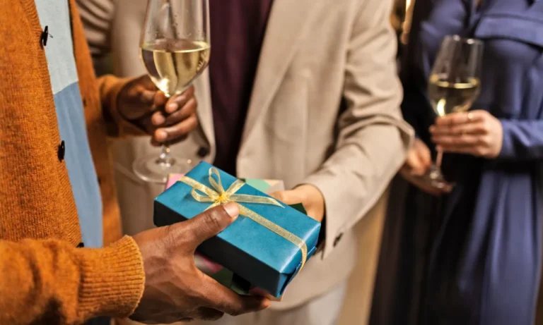 Group of people holding wine glasses and exchanging a beautifully wrapped blue gift box with a golden ribbon.