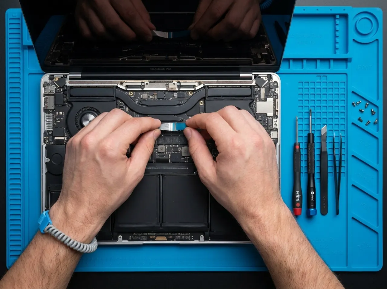 Overhead view of a technician repairing an open laptop ribbon cable on a blue anti-static mat.