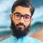 Portrait of a young man with a dark beard, wearing glasses and a light blue shirt against a blurred outdoor background.